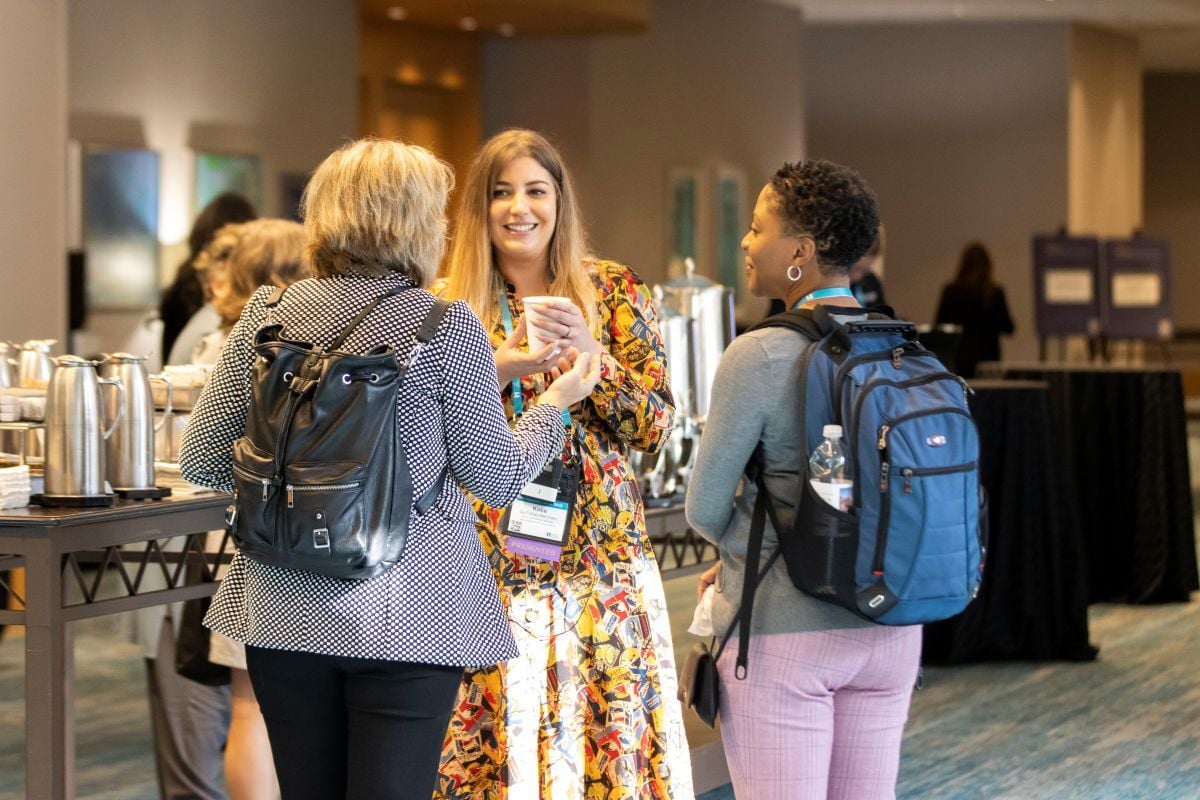 3 conference attendees talking to each other next to the conference breakfast tables.