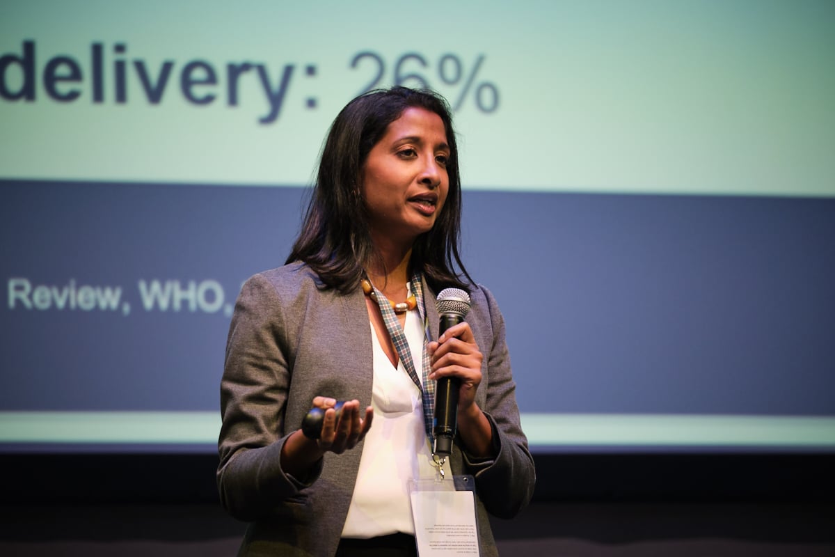 A woman with a microphone in front of a large screen giving a presentation at a conference.