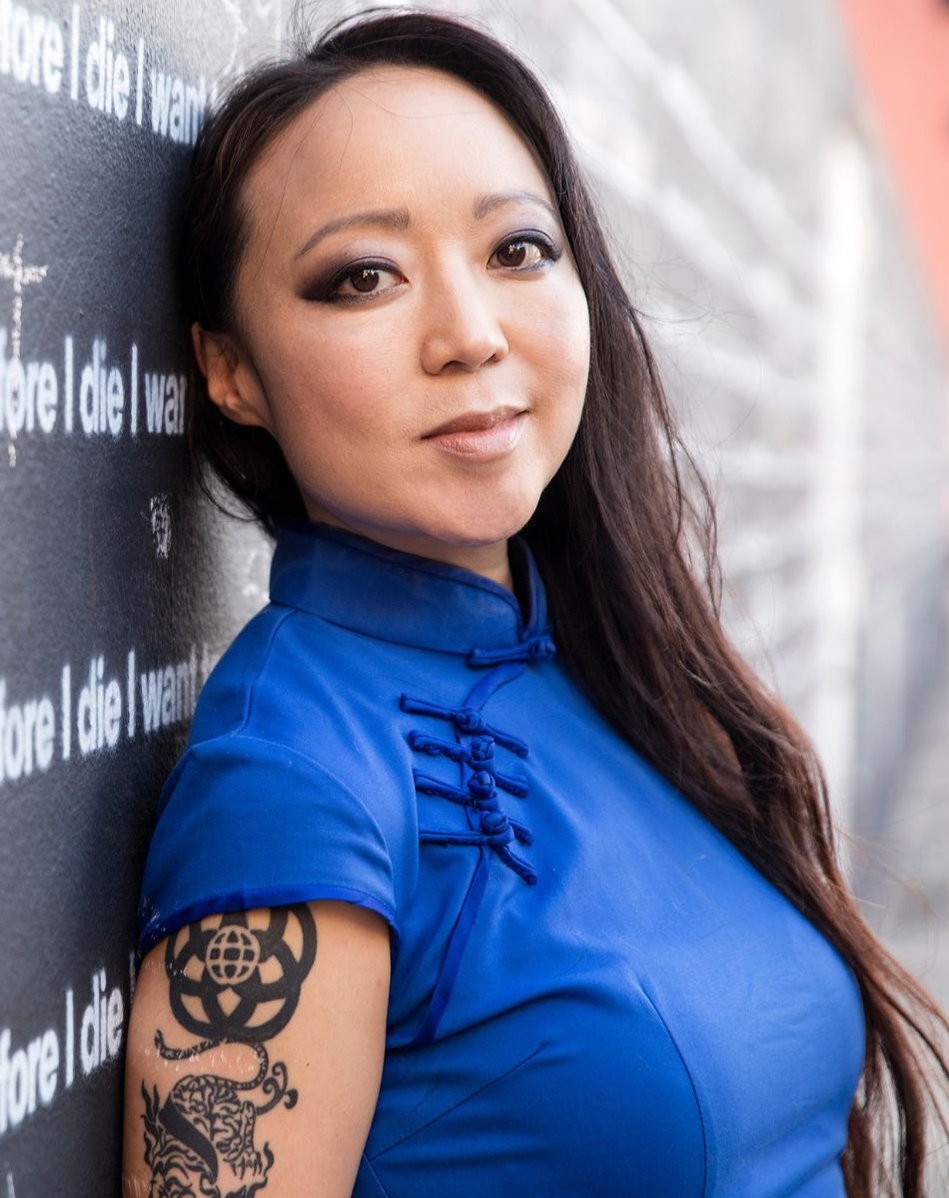 A headshot image of Candy Chang wearing a blue shirt and leaning against a black wall with white text for her