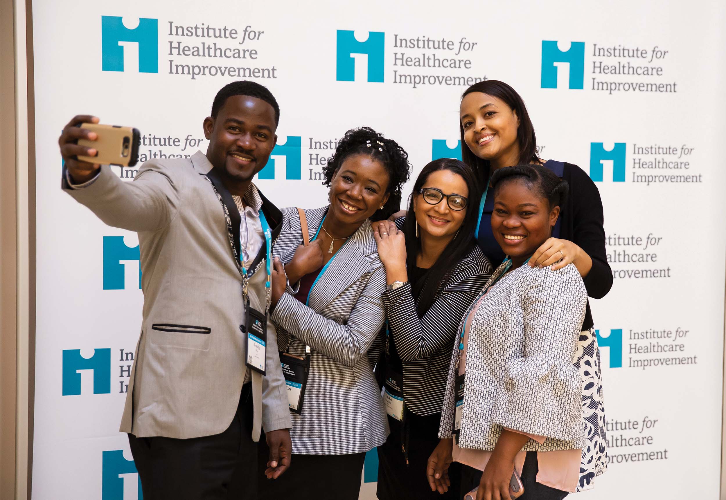 5 conference attendees taking a selfie in front of a wall of IHI logos.